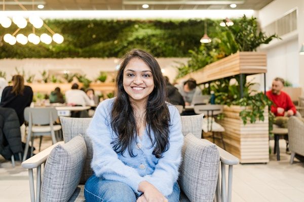 A student sat in a bright communal space with other students sat in the background and houseplants dotted around