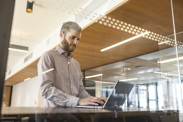 A man dressed in a shirt looks down at a laptop screen in a business environment.