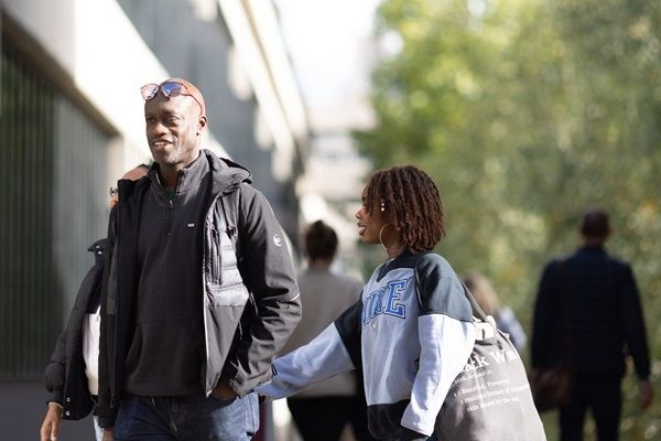 Two people walking around campus with a building and trees in the background.