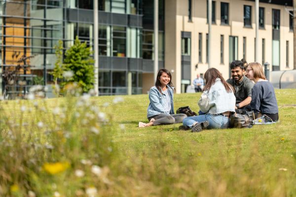 A group of four students sit cross legged on grass outside the Michael Marks building.