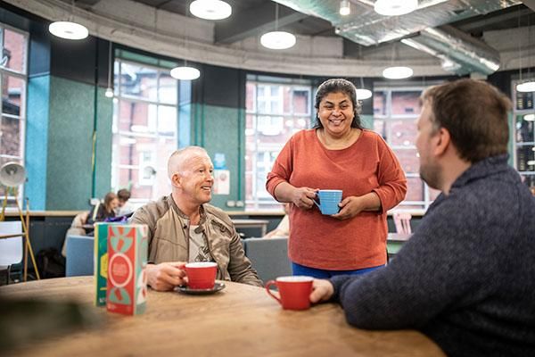 Three people chatting and drinking coffee in a cafe. Two are sitting down and one is standing up.