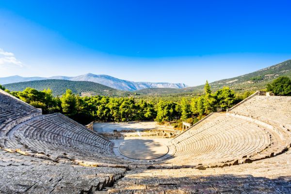 Wide view of the Ancient Theatre of Epidaurus in Greece, showing sweeping semicircular stone seating descending to a circular stage, surrounded by green forested hills and mountains under a clear blue sky.