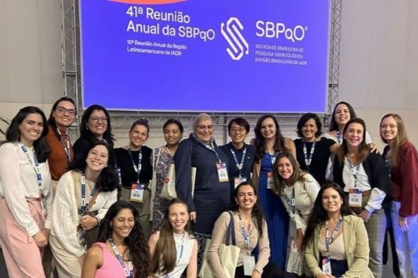 A large group of people stand together, smiling at the camera. They all have lanyards around their necks and behind them is a screen with text in Portuguese.