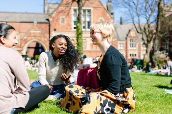 Three smiling students sitting on the grass in front of a redbrick building on campus.