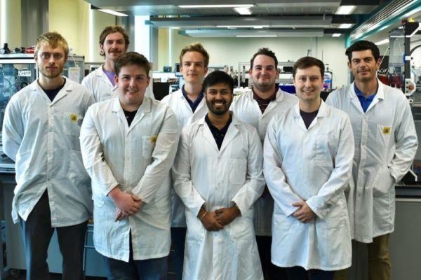 A group of people wearing white lab coats standing together in a laboratory, with scientific equipment and workstations visible in the background.
