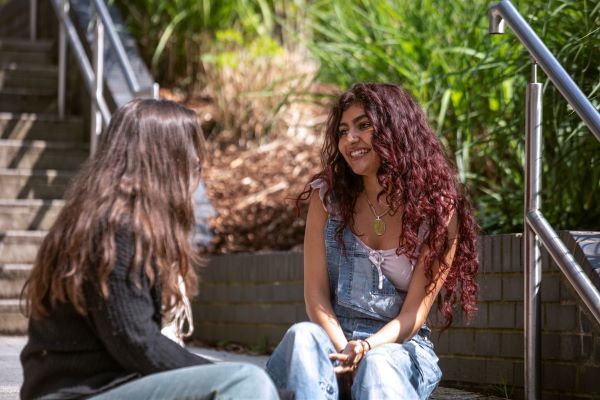 Two students sit on a set of stairs outside, facing each other and talking.