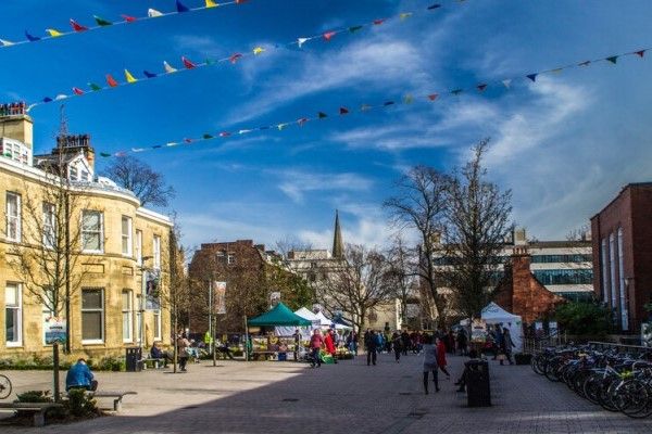 Outside Leeds University Union: an open pedestrian space with buildings on either side and colourful bunting overhead. People are walking around and visiting stalls in gazebos.