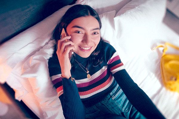 A prospective student sits on their bed smiling past the camera with their phone to their ear.