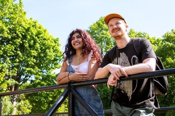 Two students stand next to each other, leaning on a metal bridge. They smile into the distance, against a backdrop of green trees and blue sky.