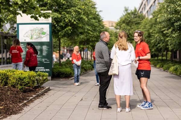 A student ambassador in a red T-shirt helps visitors on campus, surrounded by families and greenery.