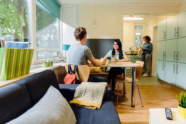 Two students sit at a table in a lounge and a third student stands in a kitchen drying cutlery.