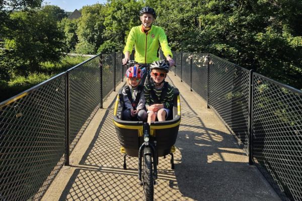 Two children wearing helmets sit in the front cargo area of a black electric cargo bike while an adult in a high-visibility jacket cycles behind them across a narrow metal bridge, surrounded by dense green trees in bright sunlight.
