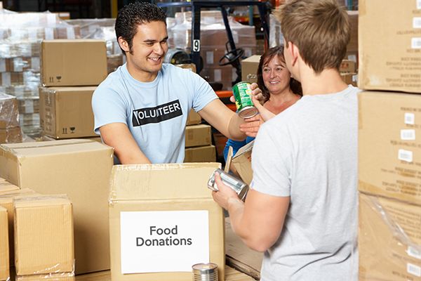Three volunteers in a warehouse, surrounded by cardboard boxes. One box says 'food donations' on it. One volunteer hands another a food can.
