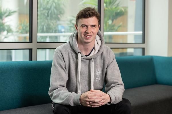 A student wearing a grey University of Leeds hoodie sitting with their hands together on a sofa