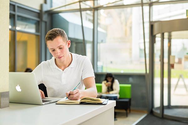 A student sat at a desk using a laptop and referring to a book.