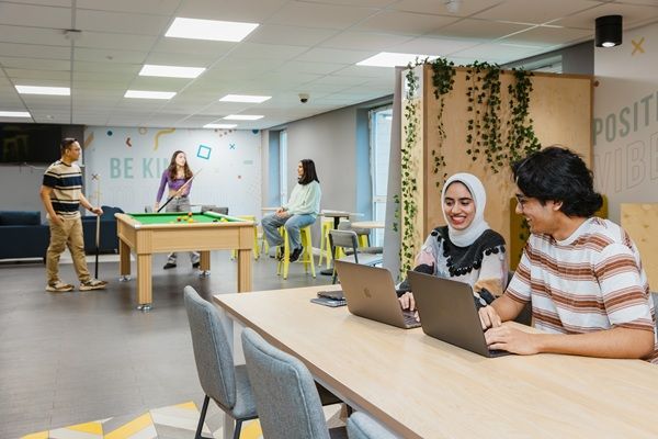 Students studying and socialising in a communal living space. Two are sat at a table using laptops and a small group are playing pool in the background.
