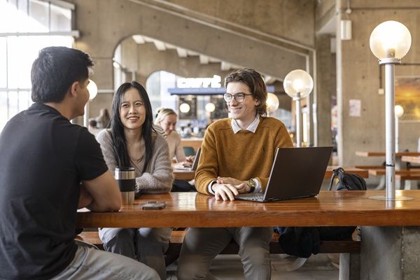 Three students sat at a table in the Roger Stevens Cafe, chatting and using a laptop