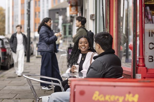 Students on the street outside a bakery chatting. Two are sat on chairs drinking hot drinks, two are stood up.