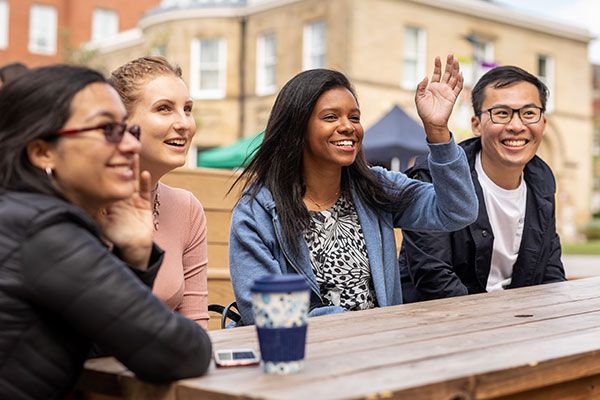 Four international students sitting at a picnic bench on campus. They are smiling and one student is waving.