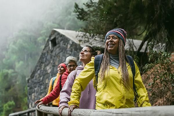 Four women look out from behind a railed walkway while trekking in a mountainous area. Behind them is a stone building, and a tree-lined slope disappearing into fog.