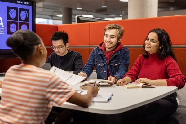 Four students sit around a table smiling and working on laptops