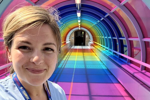 Lydia Spence wearing healthcare uniform stood in front of a tunnel-shaped walkway in the colours of the rainbow
