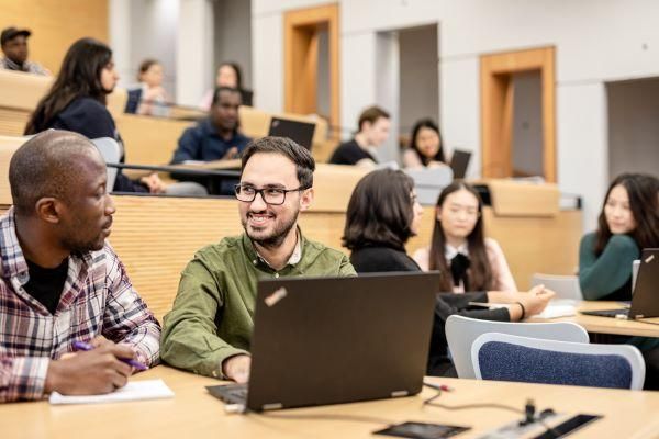 Side, front view of students in a lecture theatre. Two students chat to each other with an open laptop in front of one and the other is holding a pen with paper pad. Several other students sit behind in tiered seating.
