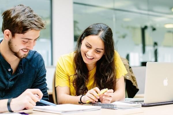 Two students sat at a desk, chatting and smiling. there is a laptop and documents on the desk.