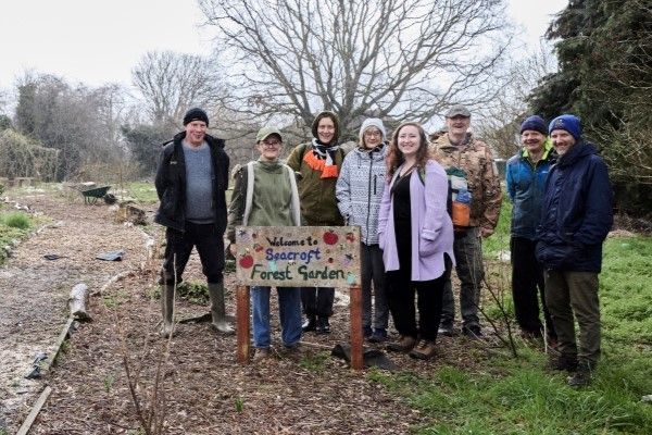Group of people at Seacroft Community Forest Garden standing with a sign reading 'Welcome to Seacroft Forest Garden', including community members and University of Leeds researchers.
