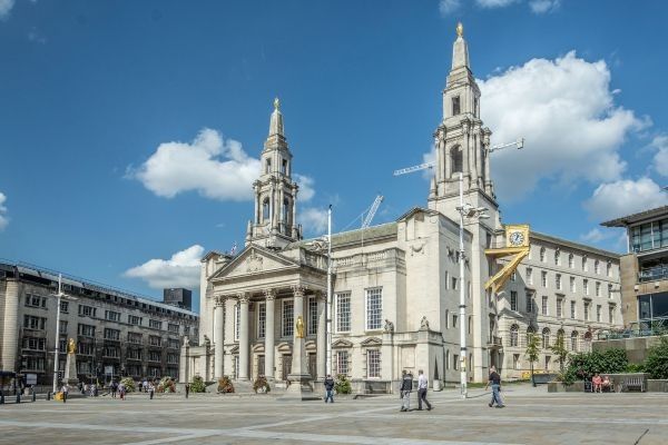A grand stone civic building with tall twin clock towers, large arched windows, and a columned entrance stands under a bright blue sky with scattered clouds. The open square in front of the building has a few people walking across wide paved area.