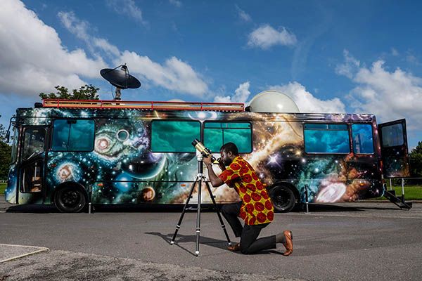 A bus decorated with space and planet imagery, with a person using a telescope in the foreground.