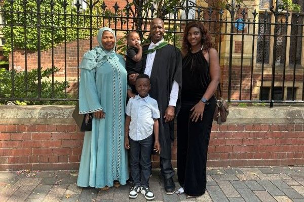 Musa Cham smiles outside the Great Hall at the University of Leeds, wearing his graduation robes and carrying his baby daughter as his sister, wife and son stand around him.