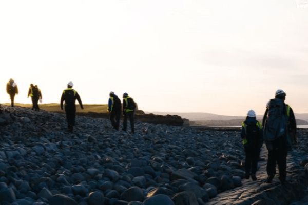 People walk away from the camera over a large pile of rocks and rubble, wearing protective equipment.