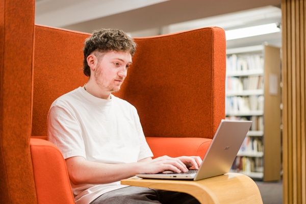 A student sat in a bright orange study booth in a library, using their laptop.