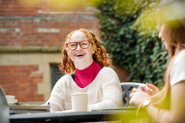 Two students sat at a table outside on campus with hot drinks. One is laughing.