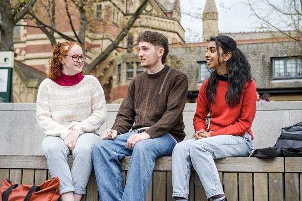 Three students sat outside on campus on a winters day, with the Great Hall behind them, chatting