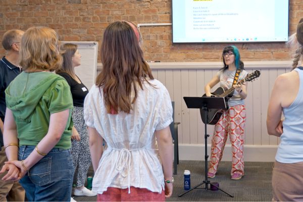 A collage showing (left): a group of people practising for a choir performance; (right) Sue Jennings and Anna Douglas in full costume as part of their research. Sue wears a pink woopee cushion costume and Anna wears a yellow pineapple costume.