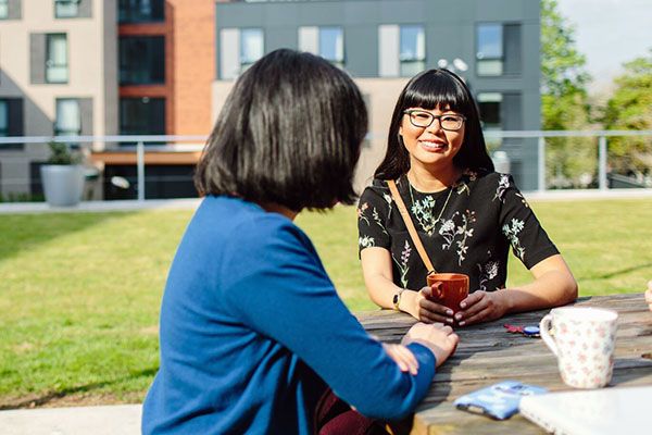 A student pictured sitting at a picnic table and talking with a friend at St Marks Residences, on a sunny day.