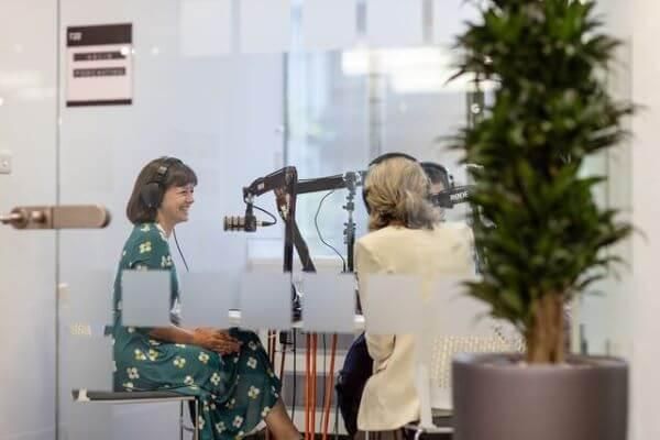 People sit in a studio in Helix around a table wearing headphones and speaking into microphones, One person is sat side on in full view and the other two sit facing away from the camera.
