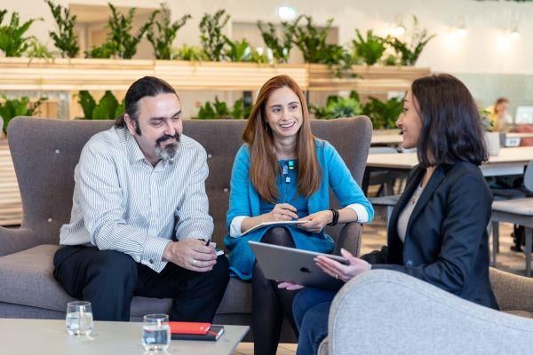 Three people sit on sofas smiling and chatting. One person to the right is showing the other two a tablet screen. There are notebooks on a low table next to them.