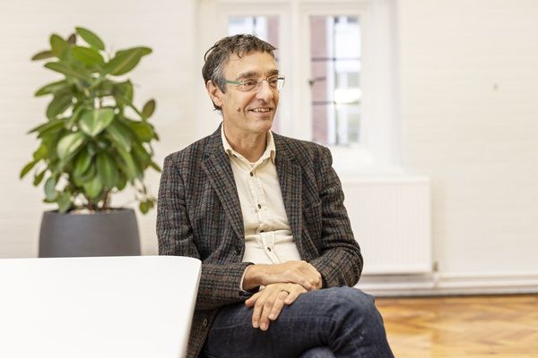 Man with glasses grey blazer sits in front of window and green indoor plant.