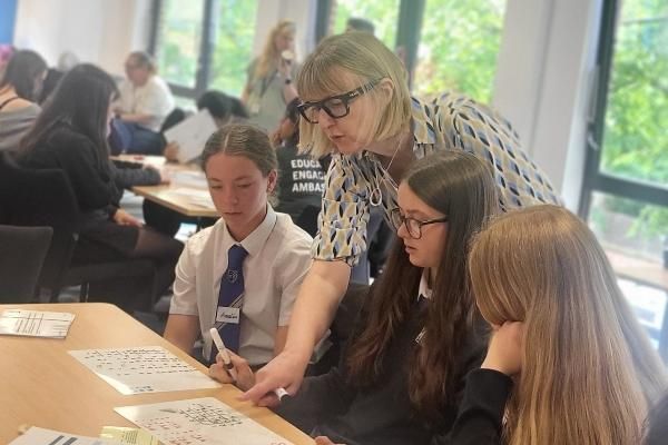 A woman wearing glasses is leaning down at a desk and instructing three female students.