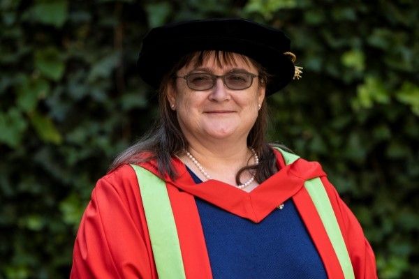 Rachel Lord stands smiling at the camera wearing red and green graduation robes and a black cap.