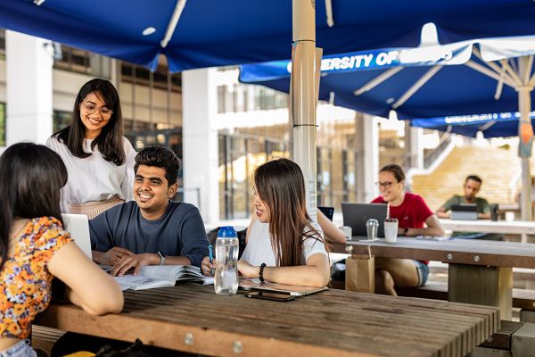 Three students sat at a picnic bench on campus doing work. A fourth student is stood next to the bench and more students are working in the background.