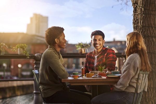 Student Rohan Cheerala smiling, sat at a cafe table with friends. The Leeds skyline is in the background