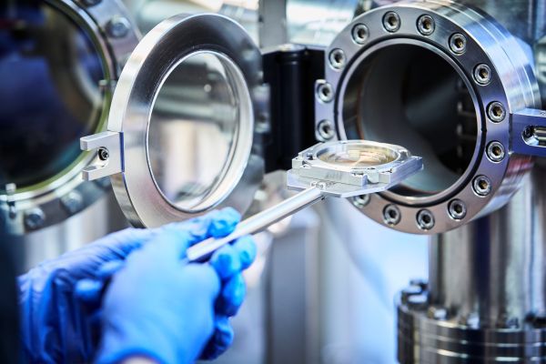 Blue gloved hands position a circular sample holder inside into the System’s open high-vacuum deposition chamber, a circular mental opening. 