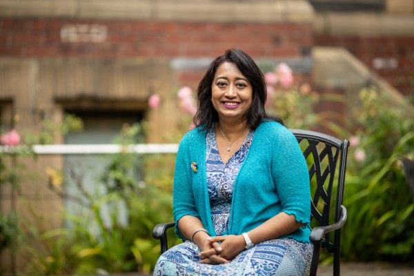 Rumana Hossain in chair outside, in front of plants and a red-brick wall.