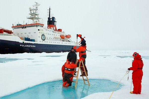Three people doing tests in some water that has appeared in a hole on an ice sheet in a frozen environment. A large boat is in the background.
