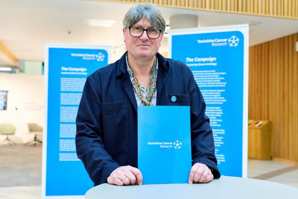 Man with glasses stands in front of blue banner and holds blue folder that reads Yorkshire Cancer Research