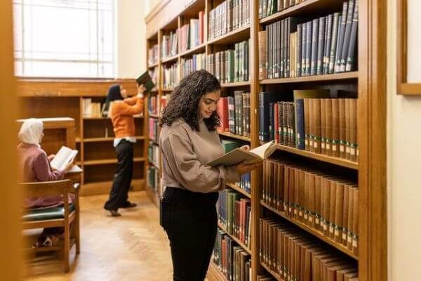 A student stands at a wooden bookshelf reading an open book. They are wearing a brown long sleeved top and dark trousers.